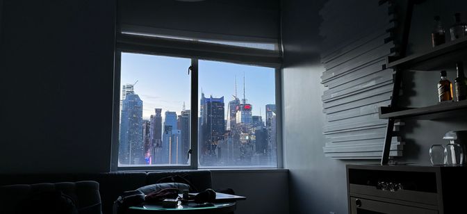 Photo from a bed indoors. Large window. Sunrise. Skyscrapers in the distance.